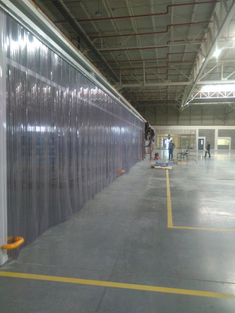 Industrial warehouse interior with transparent plastic partitions, yellow floor markings, and workers in the distance under bright overhead lighting